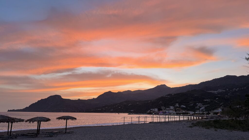 Kreta, Blick aus einer Bucht vom Strand Richtung Berge, darüber ein orange-grauer Himmel