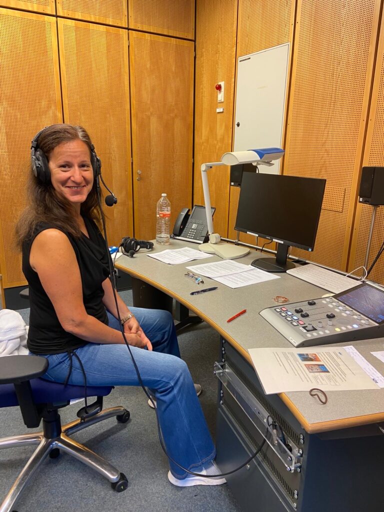 Astrid Schmidt sitzt gut gelaunt in einem Radiostudio an einem Arbeitsplatz mit Kopfhörer und viel Computertechnik auf dem Tisch.